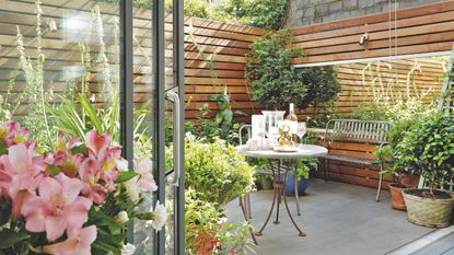 small enclosed garden with wooden cladding walls, large rectangular mirror, containers and pots, crittall doors, iron bench, table and chairs 