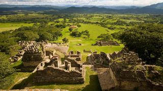 The Maya ruins of Toniná in Chiapas, Mexico. Here we see several stone buildings with their roofs and half their walls missing overlooking a beautiful green valley with trees and mountains in the distance.