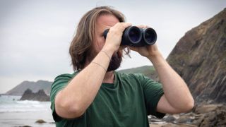A male using the Canon 18x50 IS binoculars with the sea and coastline behind them.