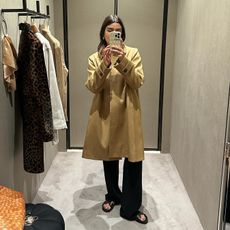 female fashion influencer Anna Newton of The Anna Edit poses for a mirror selfie in a fitting room wearing a trench coat, black pants, and flat black sandals