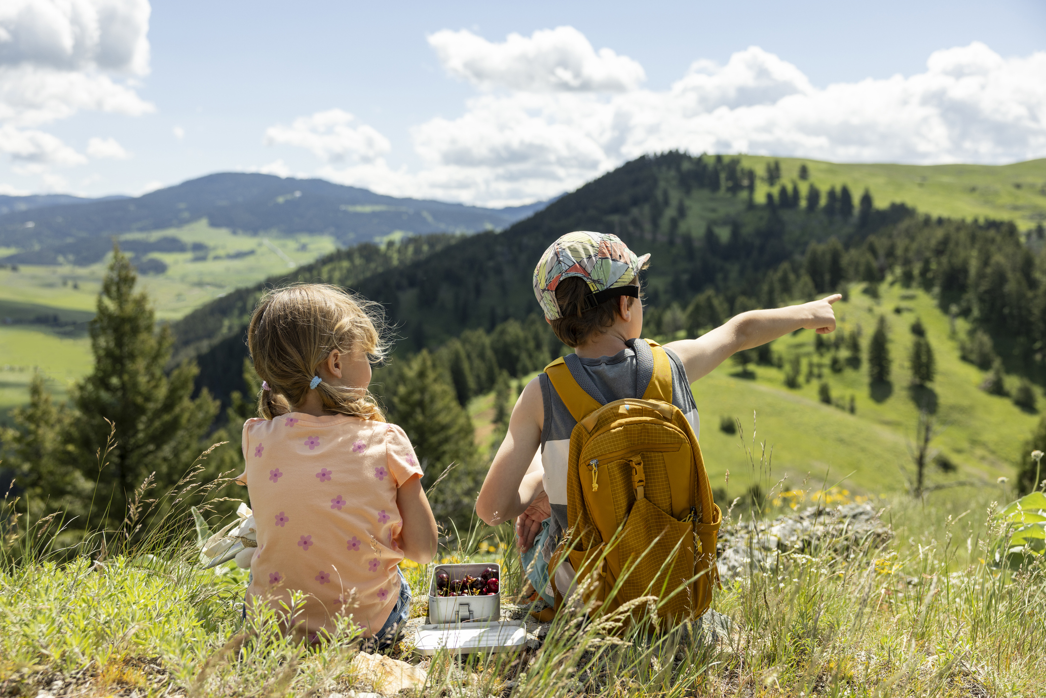 Two young children sitting on a luscious, green hill, eating lunch together.