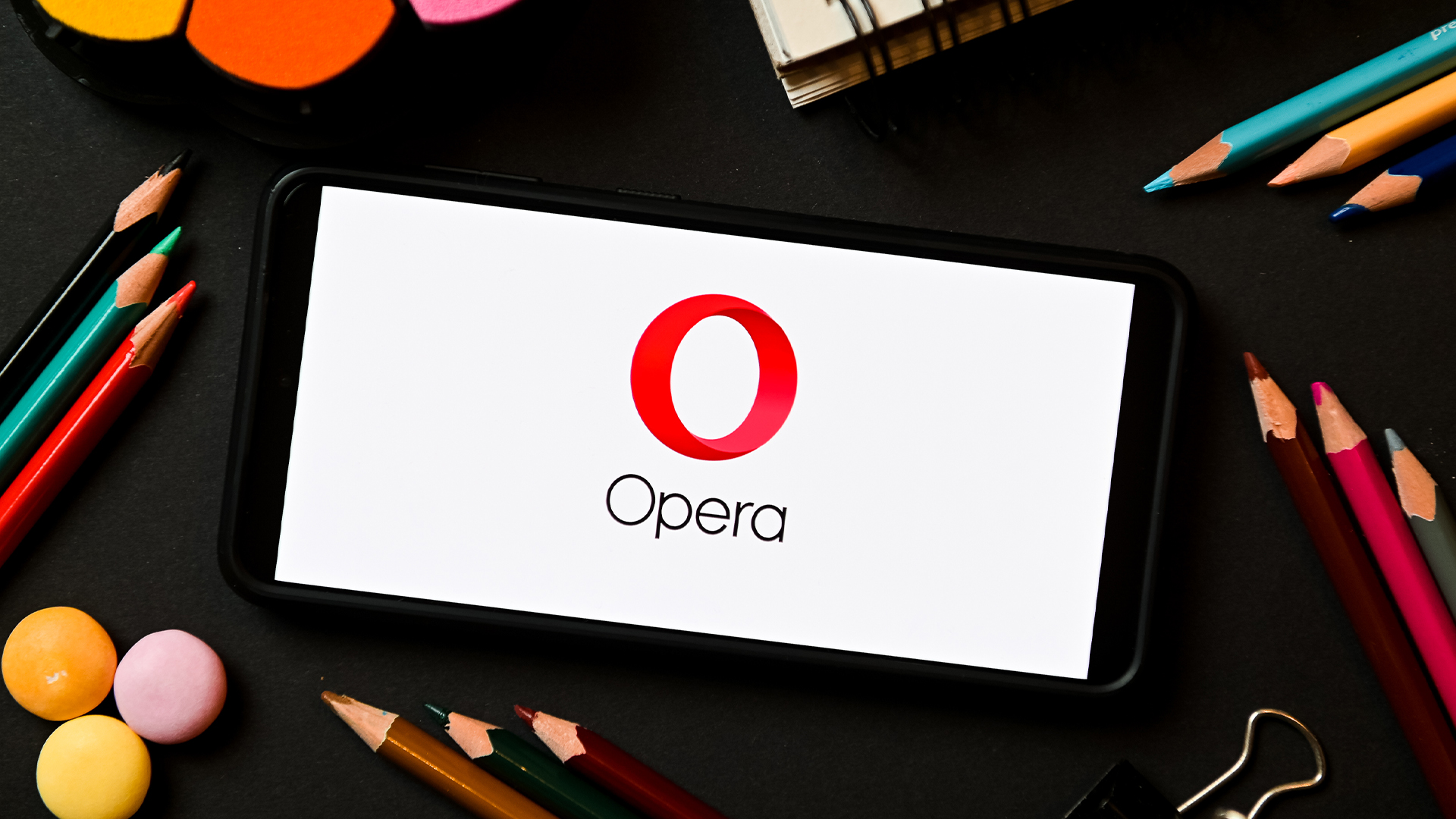 Opera browser logo and branding pictured on a smartphone screen placed on desk with pencils and art utensils.