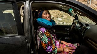 A woman sits in the passenger seat of a car, smiling, with a Sea to Summit Aeros Ultralight Traveller Pillow around her neck.