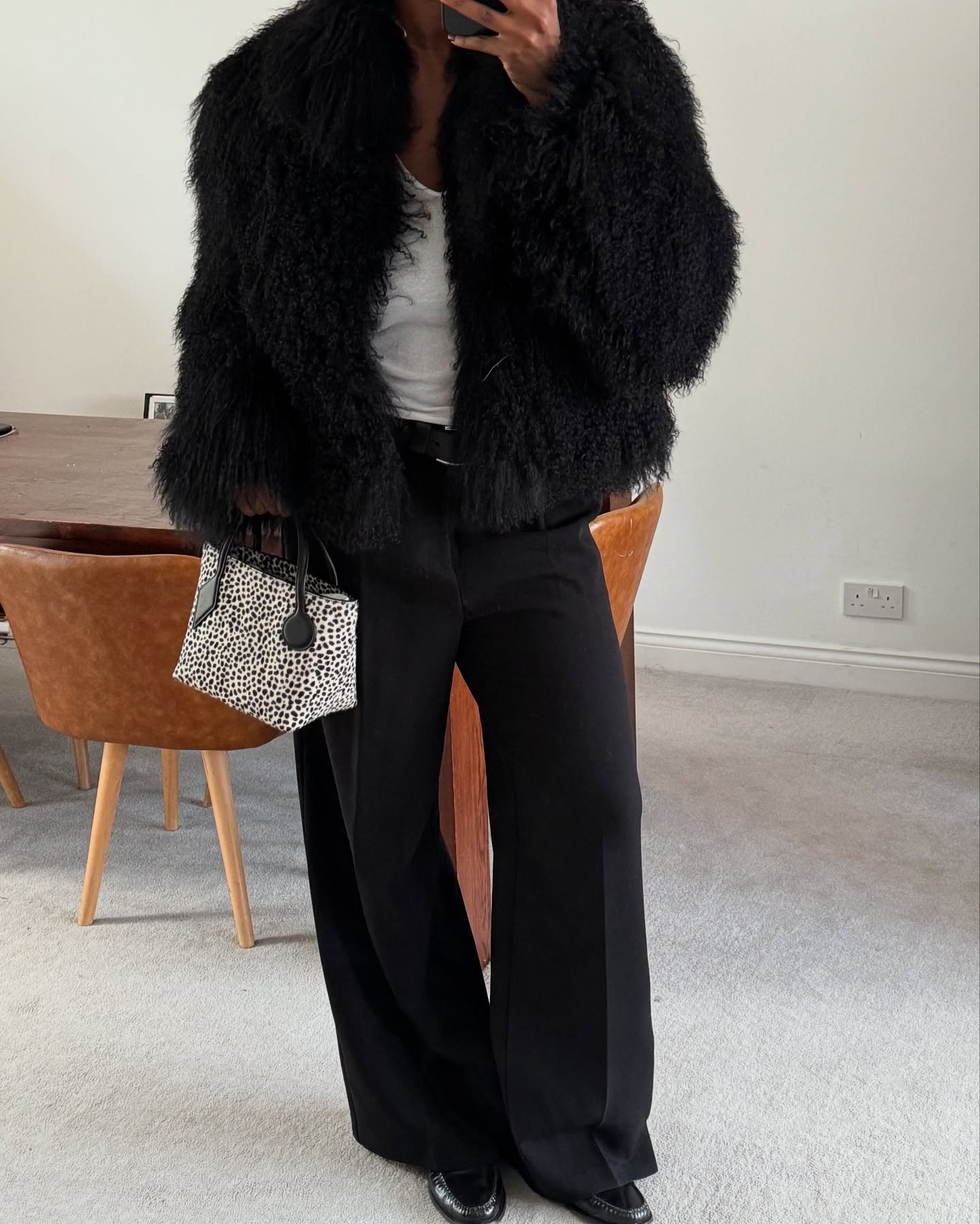 @nlmarilyn wears a black cropped fur coat, a white tee, trousers, loafers, and a dalmatian-print bag.