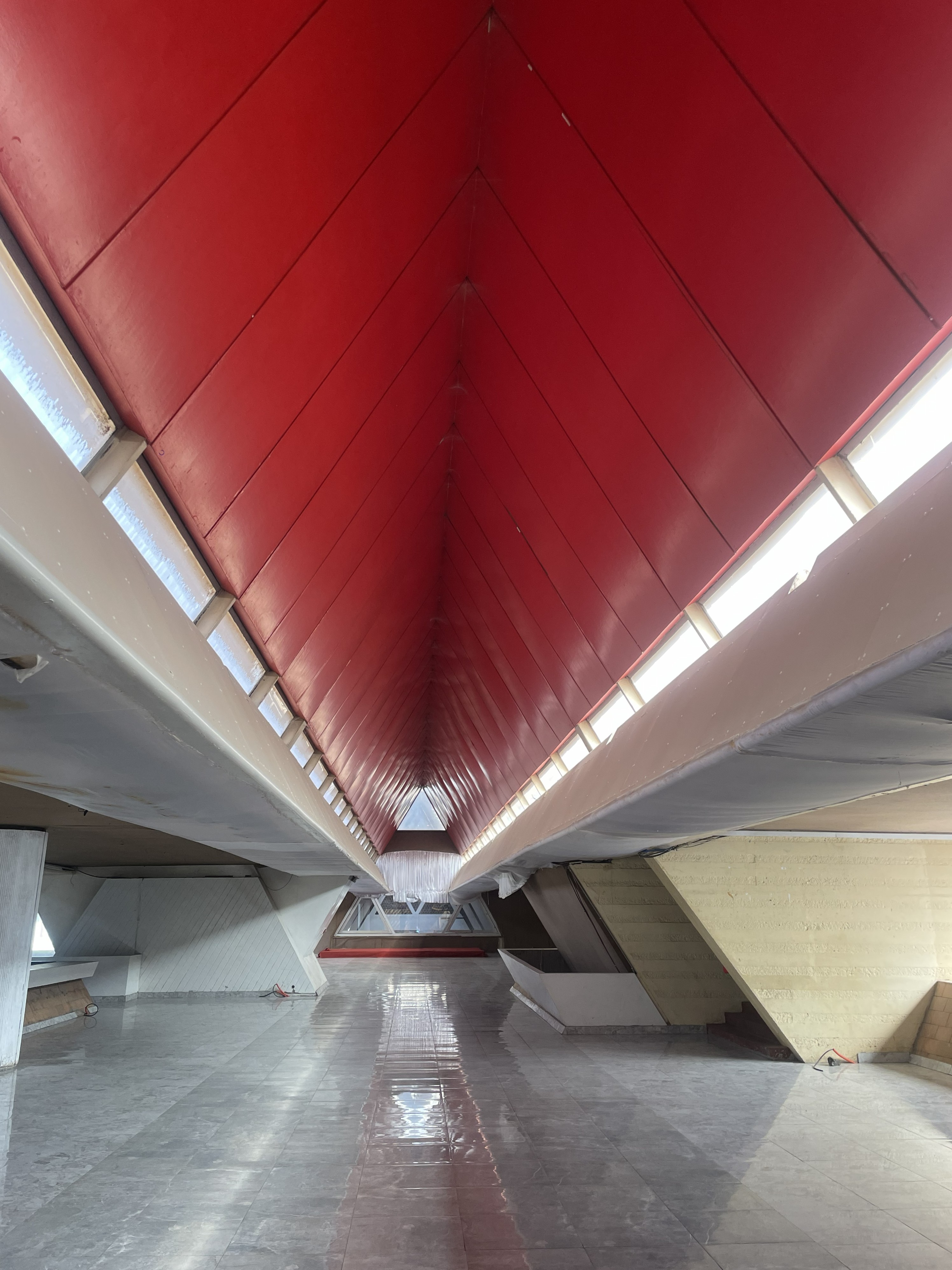 A red, lacquered tunnel with stone, reflective floors and metal-paneled red walls features a triangular roof structure, side windows, and upside-down triangular pillars.