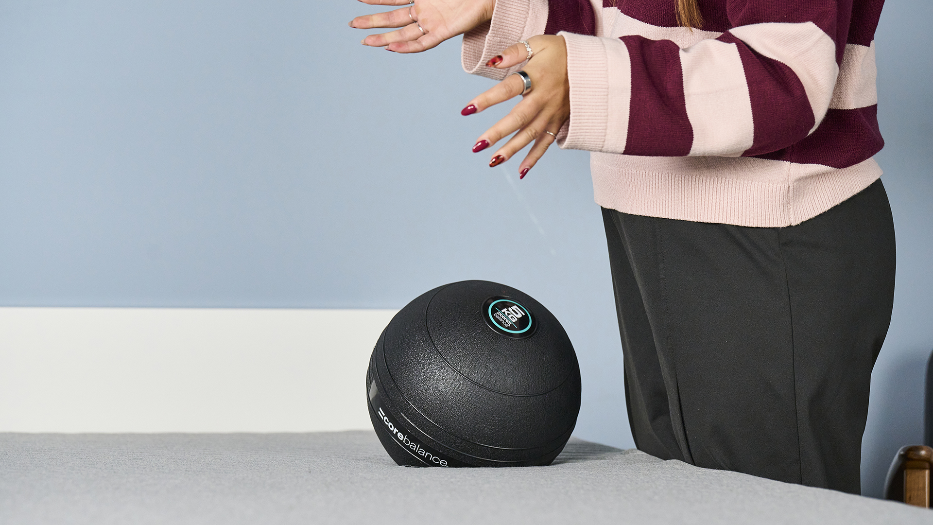 A close-up shot of a large, textured black exercise ball hitting the white surface of the Bear Elite mattress during a motion transfer test. The hands and lower torso of the mattress tester in a striped sweater who has just dropped the ball are visible on the right hand side of the bed.