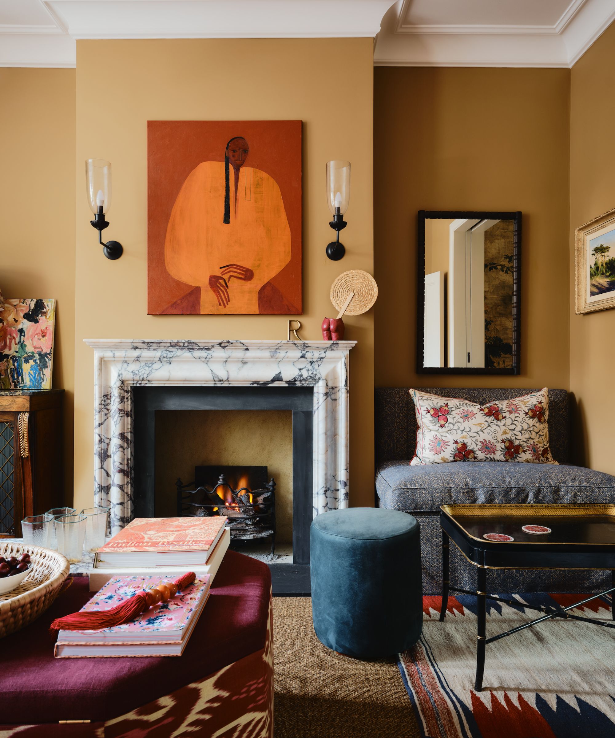 A traditional living room with dark ochre walls, a marble fireplace with a red and orange modern artwork above. Carpeted floors with layered patterned rugs, a blue loveseat, a burgundy ottoman, and a teal footstool.