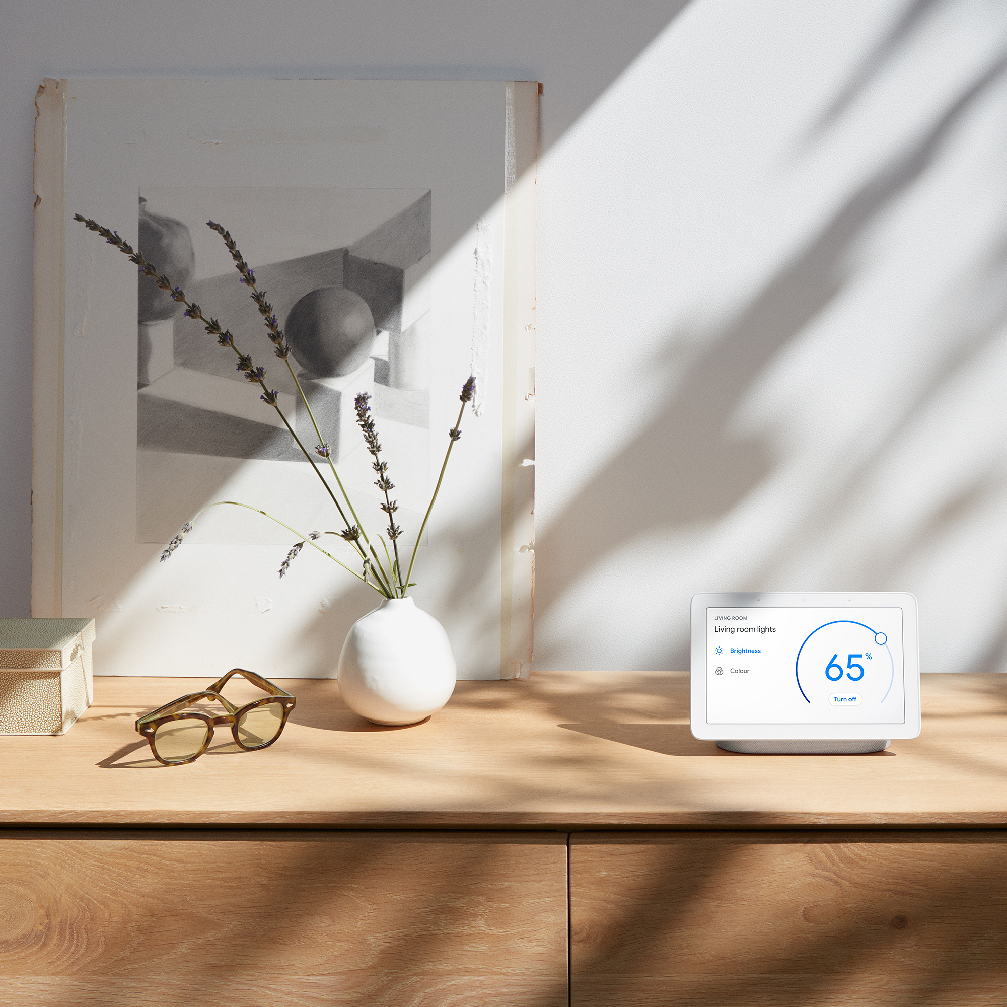 Google Home Hub on a wooden sideboard with various items like a vase with lavender springs, and framed art print and a pair of glasses beside it
