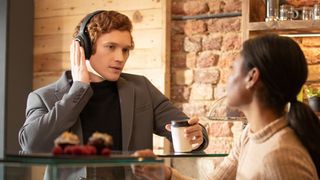 A man wearing headphones is holding a hand to his ear to activate Transparency Mode, while talking with café server