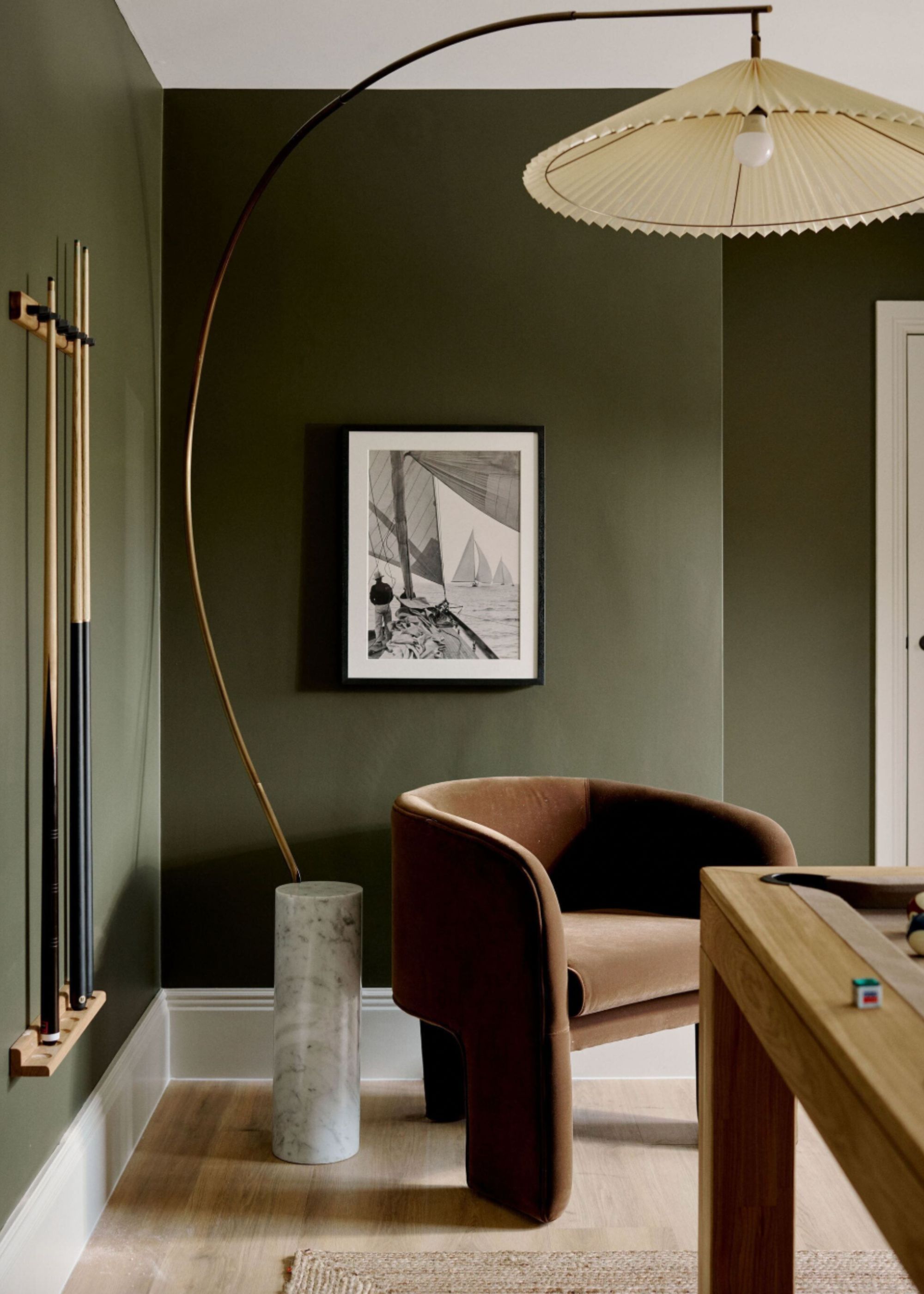 A green games room with a brown armchair and large contemporary floor lamp with a white marble cylindrical base and pleated lamp shade; pool cues are attached to the wall next to the pool table
