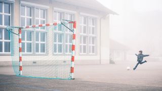 A child in a blue jacket prepares to kick a soccer ball towards a net on a foggy playground, with school building windows in the background