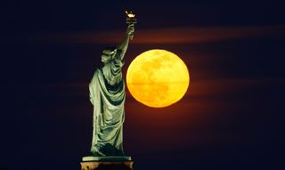 a bright, yellow-tinged full moon behind a statue of a person holding a torch aloft
