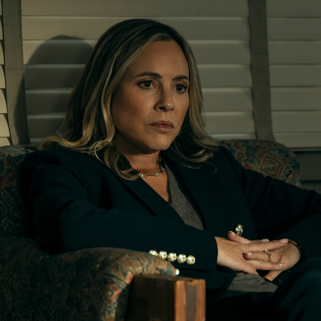 Maria Bello as Belle Buckley wearing a suit and leaning back in an office chair with her hands on her lap in the waterfront