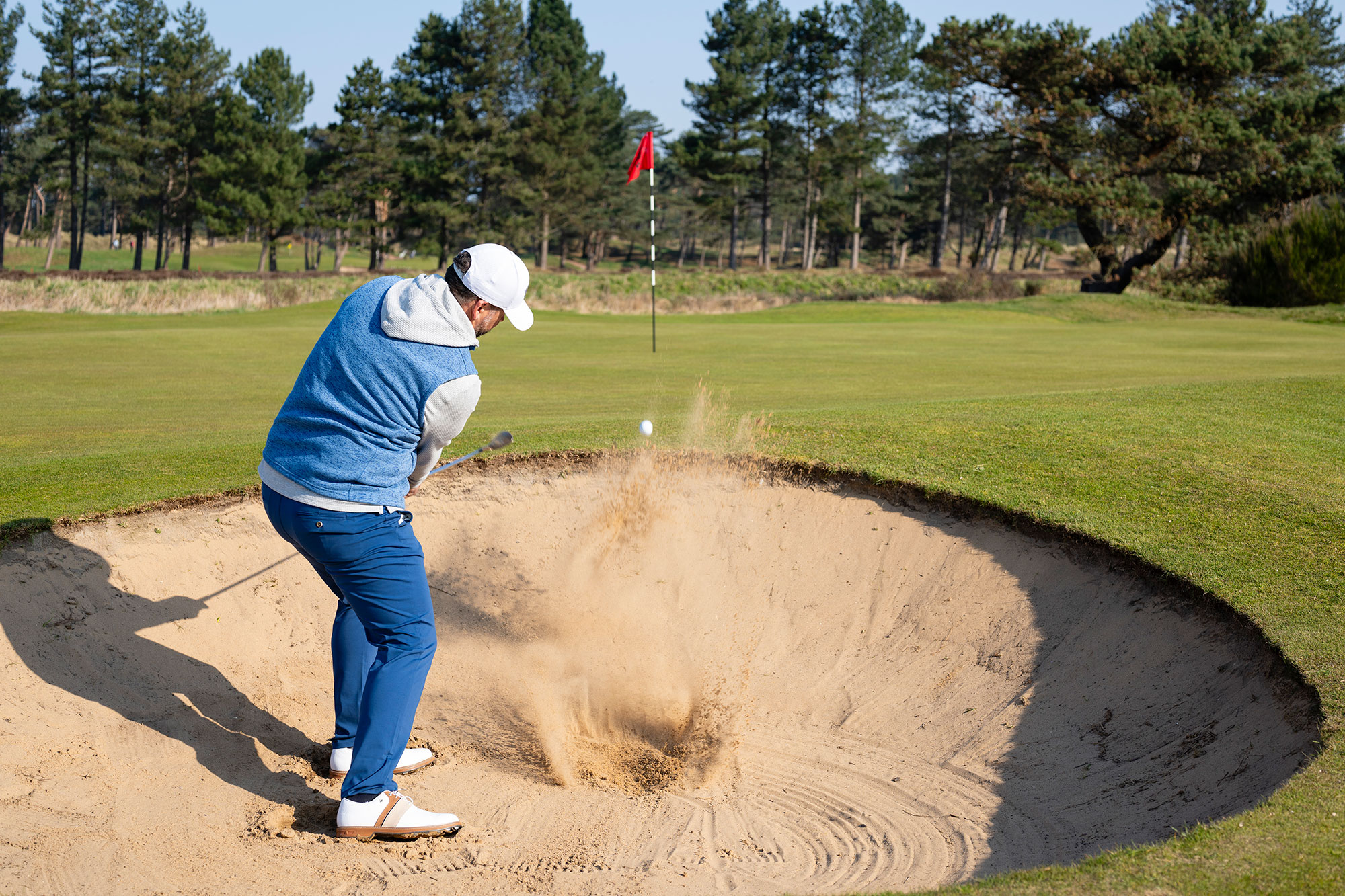 Peter Finch just past impact hitting a greenside bunker shot, with the ball propelled towards the green and an explosion of sand