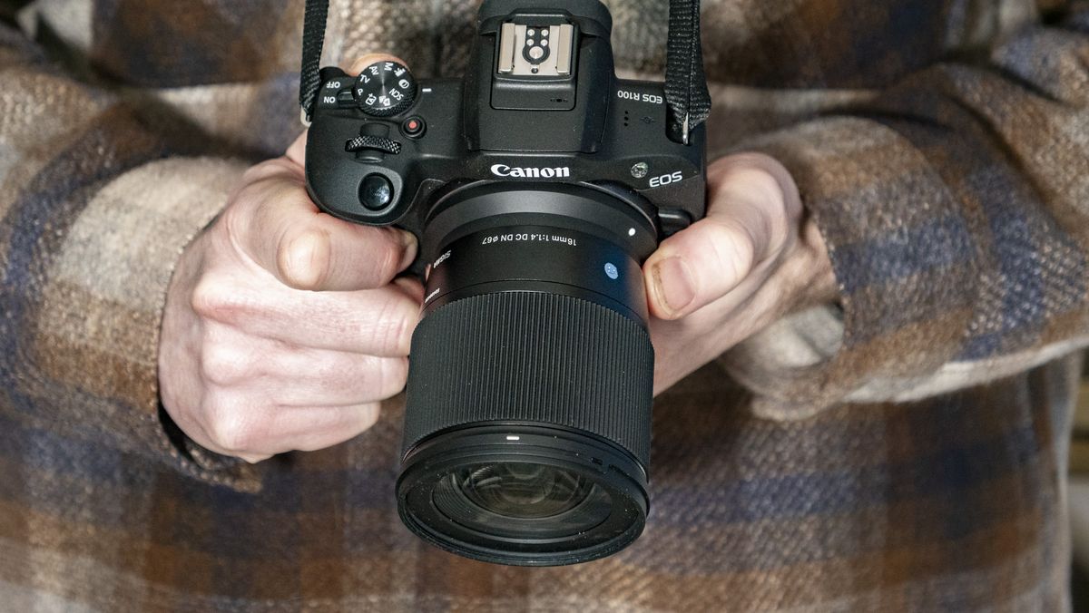 Sigma 16mm F1.4 DC DN Contemporary lens in the hand attached to a Canon EOS R100, checked shirt background