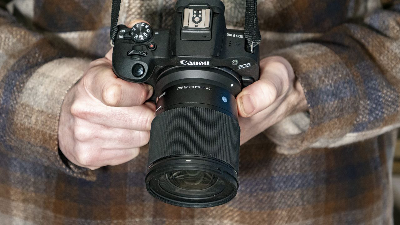Sigma 16mm F1.4 DC DN Contemporary lens in the hand attached to a Canon EOS R100, checked shirt background