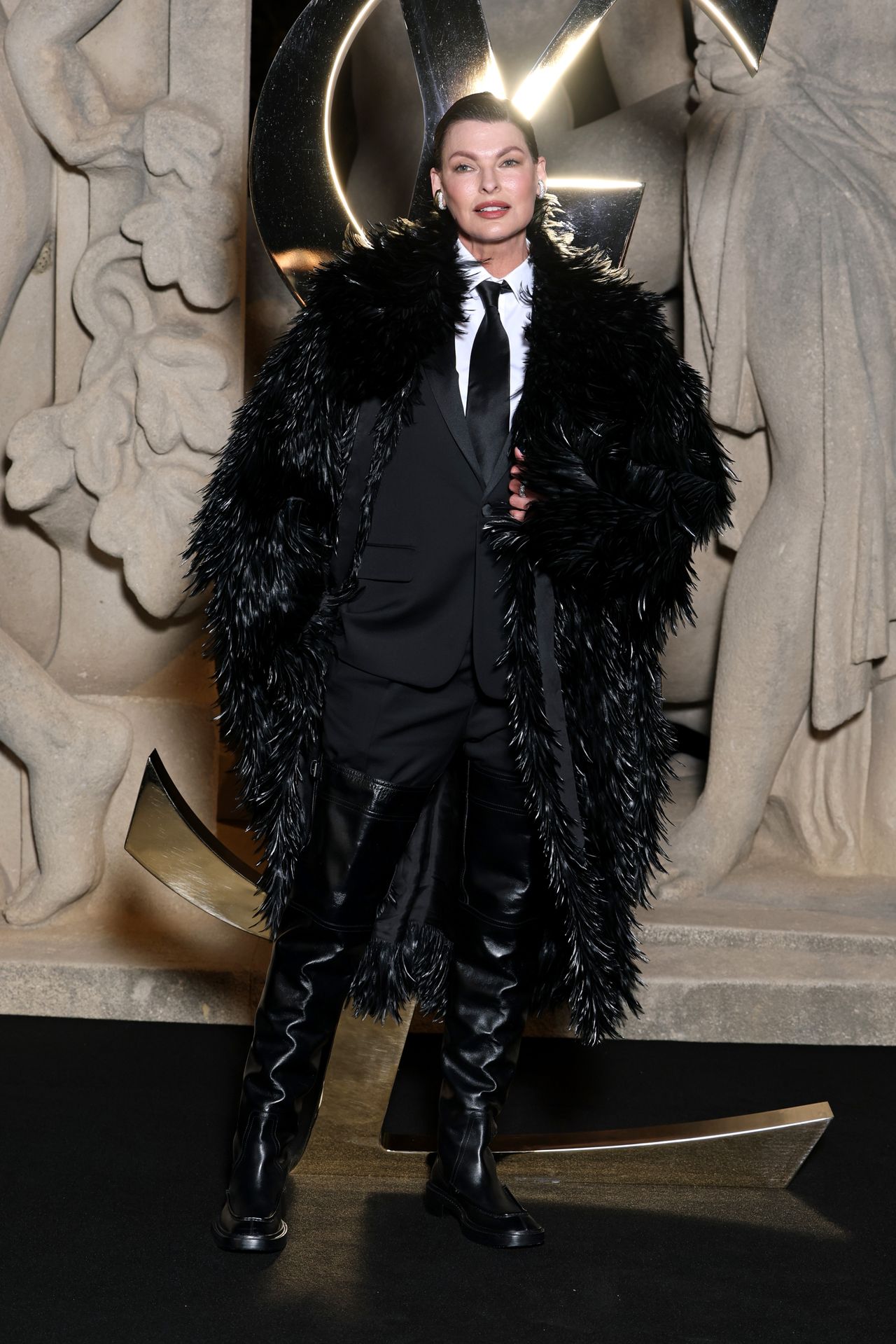 linda evangelista wearing a black feathered coat over a suit and leather boots at saint laurent