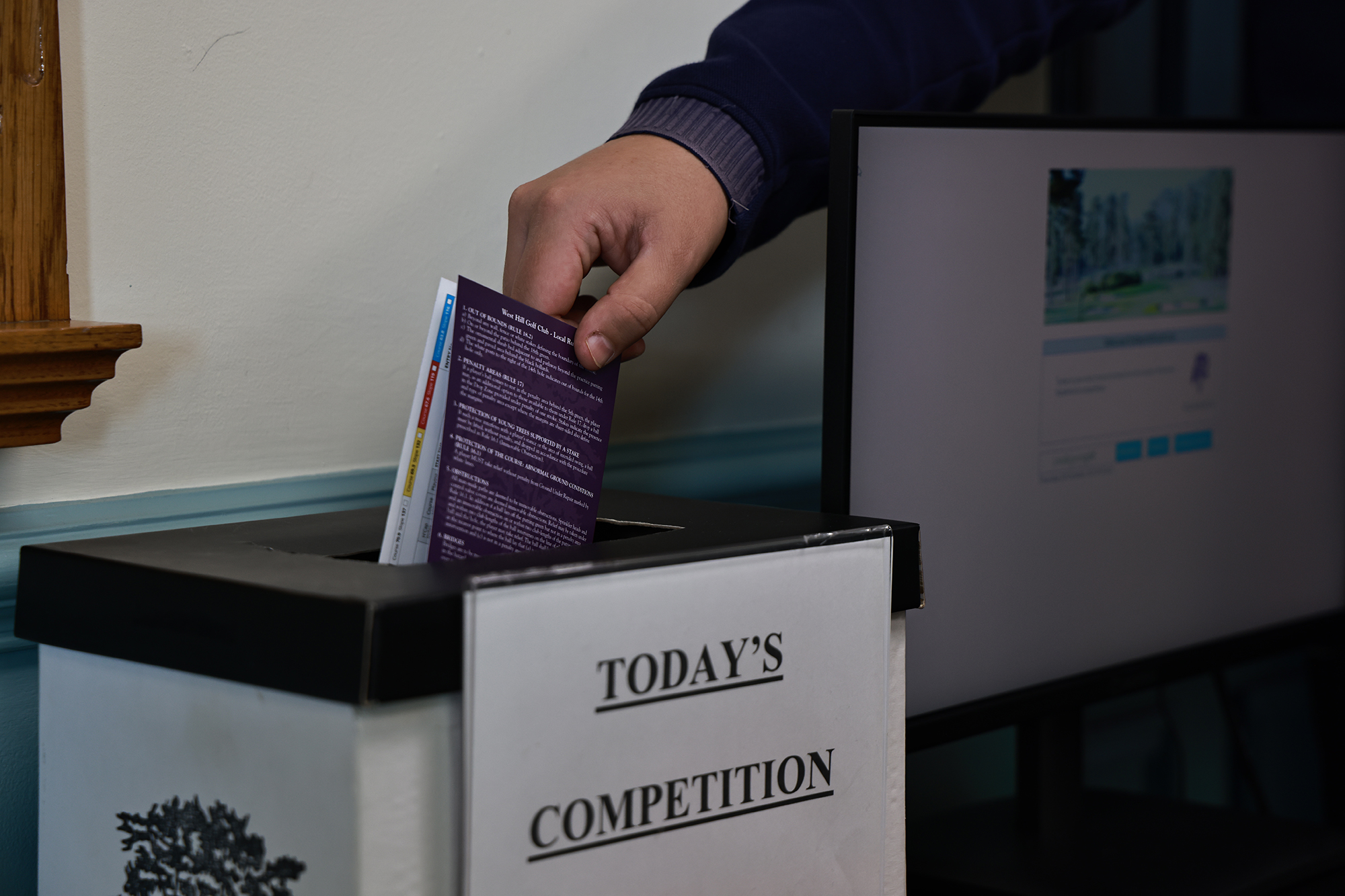 A golfer placing their scorecard into the box in the clubhouse that is labelled &#039;today&#039;s competitions&#039;, submitting their score in the process.