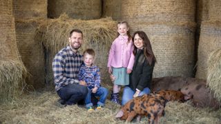 Kelvin and Liz Fletcher and their children Marnie and Milo pose together in a barn surrounded by hay bales and a pig and piglets in Fletcher's Family Farm.