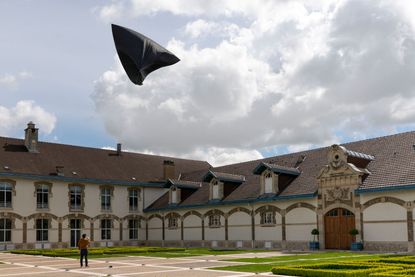 Daytime image, aeroglyphic inflated grey sculpture being flown by a person on the ground attached to rope cords, courtyard type space, paved and grass areas,, surrounding white building, brown tiled roof, windows, large wooden arched door, blue cloudy sky