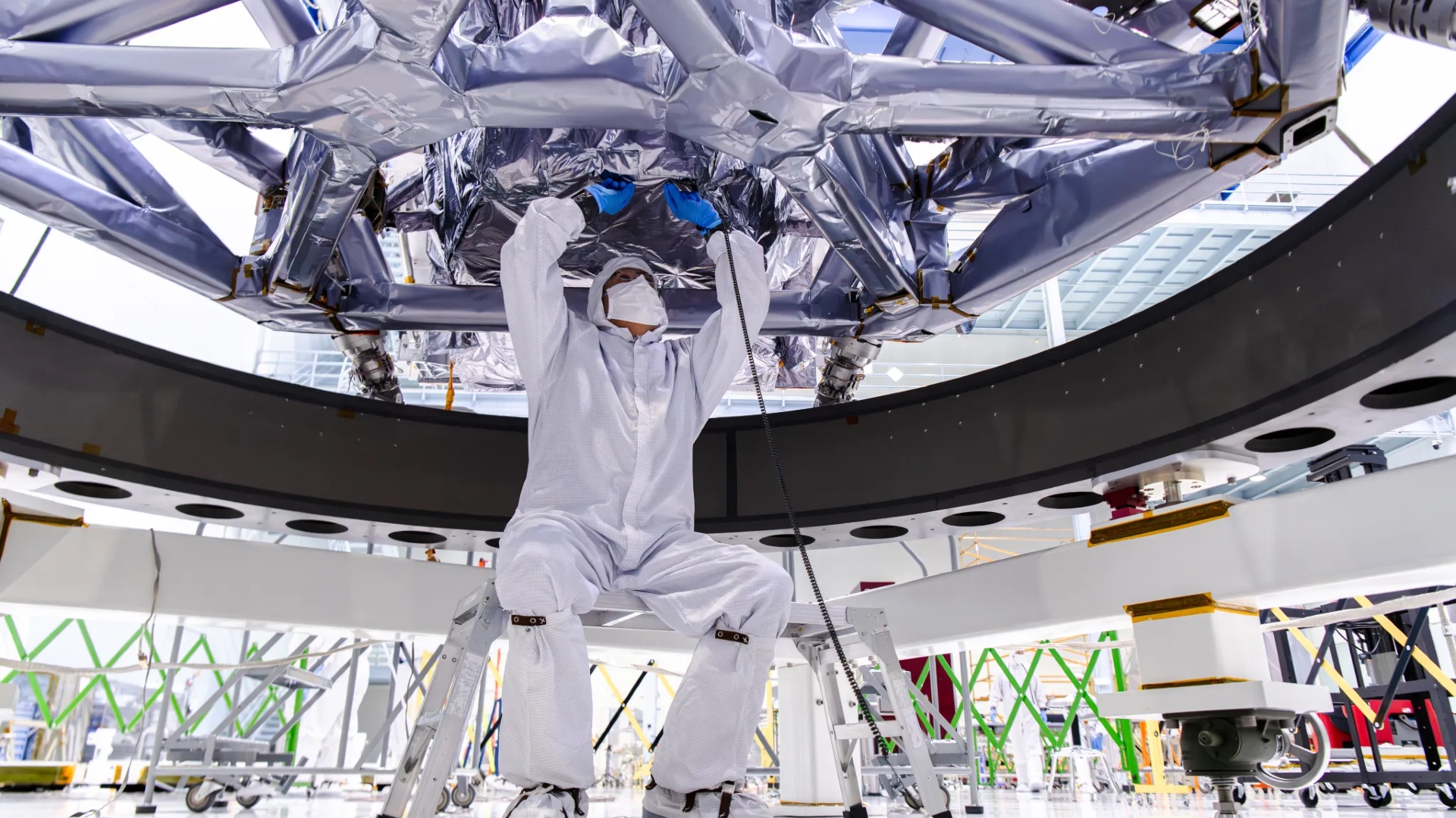 scientists in white clean suits move a metallic honeycomb-like structure onto a rectangular piece of electronics covered in wires and components