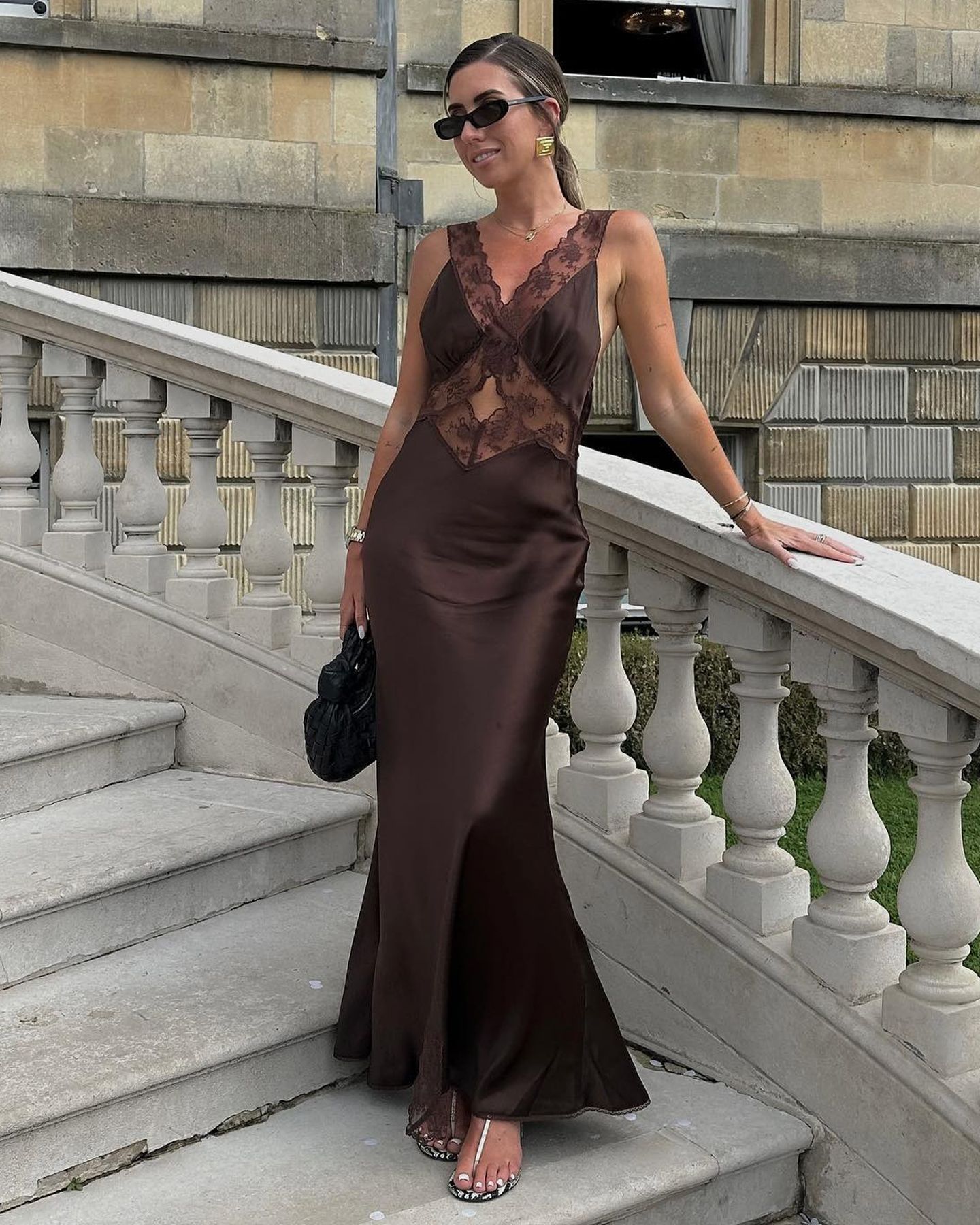 British style influencer Hannah Lewis poses at a wedding wearing rectangular sunglasses, a lace-trim black satin maxi skirt, and snakeskin sandals.