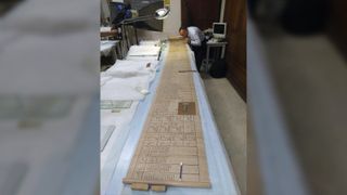 Here we see the unrolled papyrus that is the Book of the Dead spread over several meters of table in an academics office. It is being examined by a man.