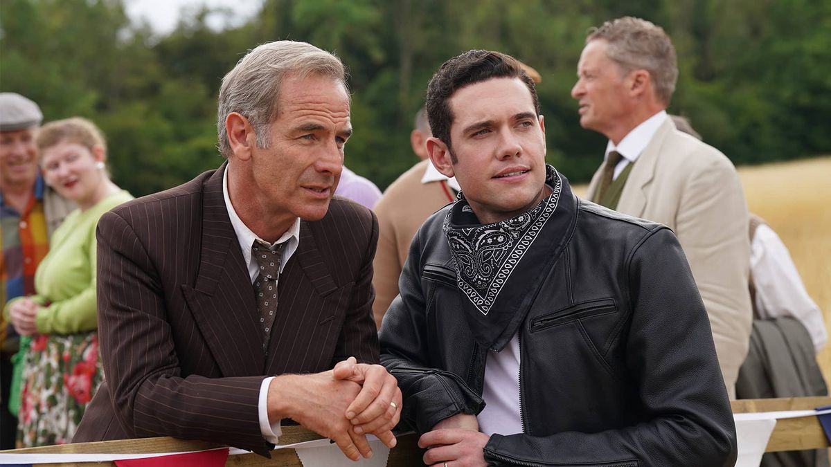 British actor Robson Green, wearing period brown suit and tie, leans against wooden gate in a scene from Grantchester season 8