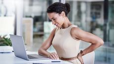 A woman sits at a desk with one hand on her laptop and one hand on her lower back. She looks upcomfortable and is grimacing