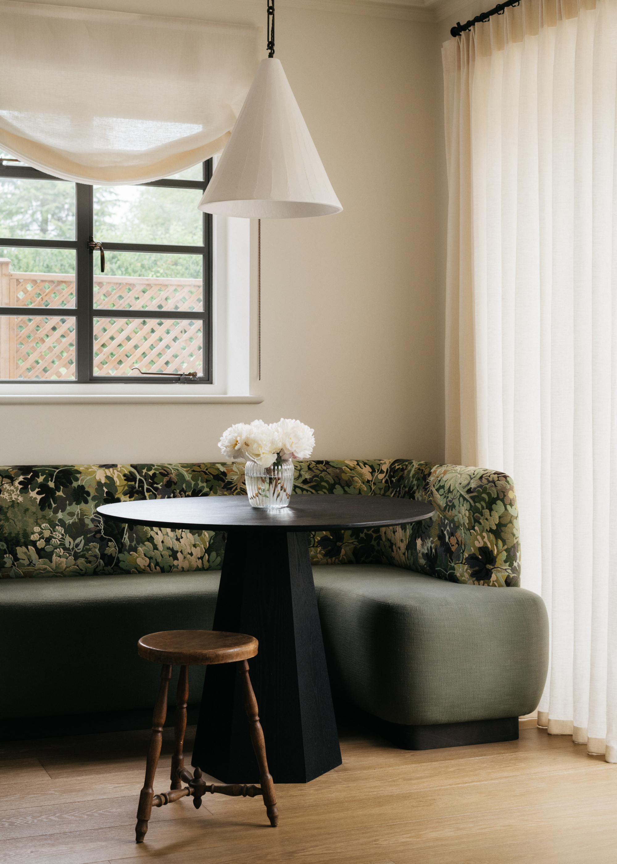 A banquette seating area with a teal base and patterned back around a round black table with a round wooden stool with flowers on the table and a pendant light above