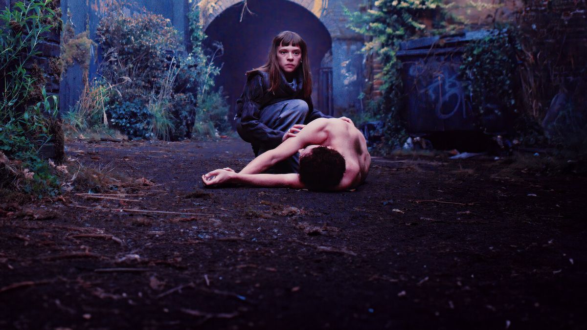 Bodies still image showing a woman standing over a body in an overgrown street