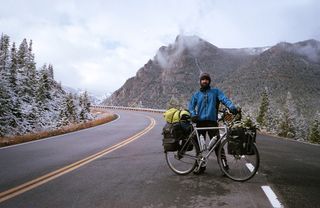 A man in winter clothing stands behind a silver steel bike, holding it upright. The bike has panniers on the front and back. In the background, snow-covered foothills of the Rocky Mountains.