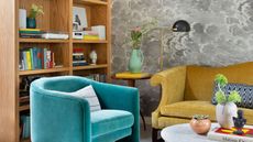 A living room corner with a light wooden bookshelf, a light blue velvet armchair, and a yellow velvet sofa. Grey cloud-like wallpaper on the wall behind. 