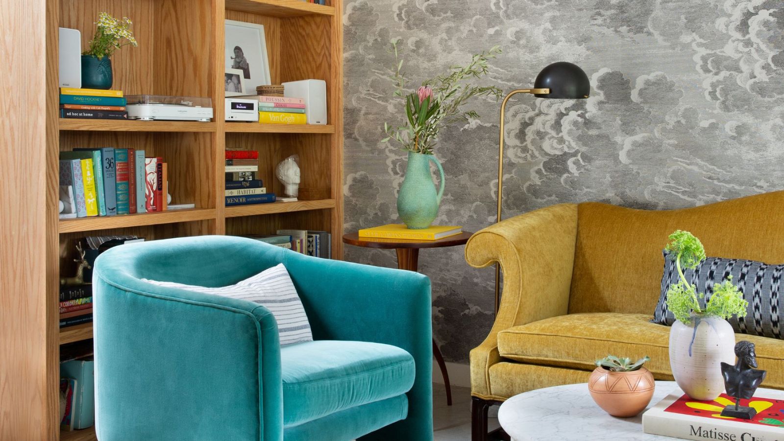 A living room corner with a light wooden bookshelf, a light blue velvet armchair, and a yellow velvet sofa. Grey cloud-like wallpaper on the wall behind. 