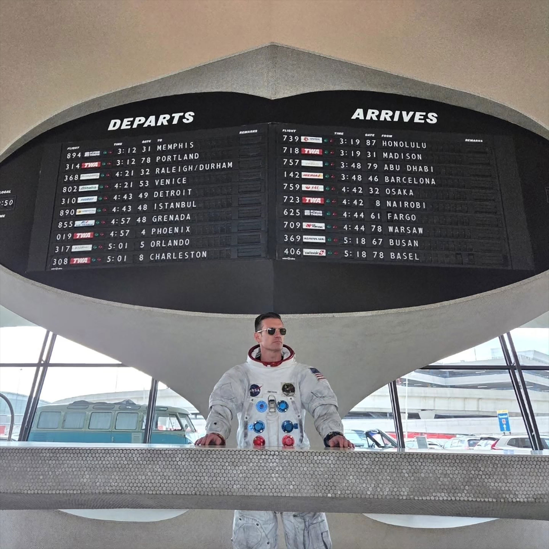 a man wearing a white spacesuit and sun glasses stands inside a retro airport terminal under a flight schedule board