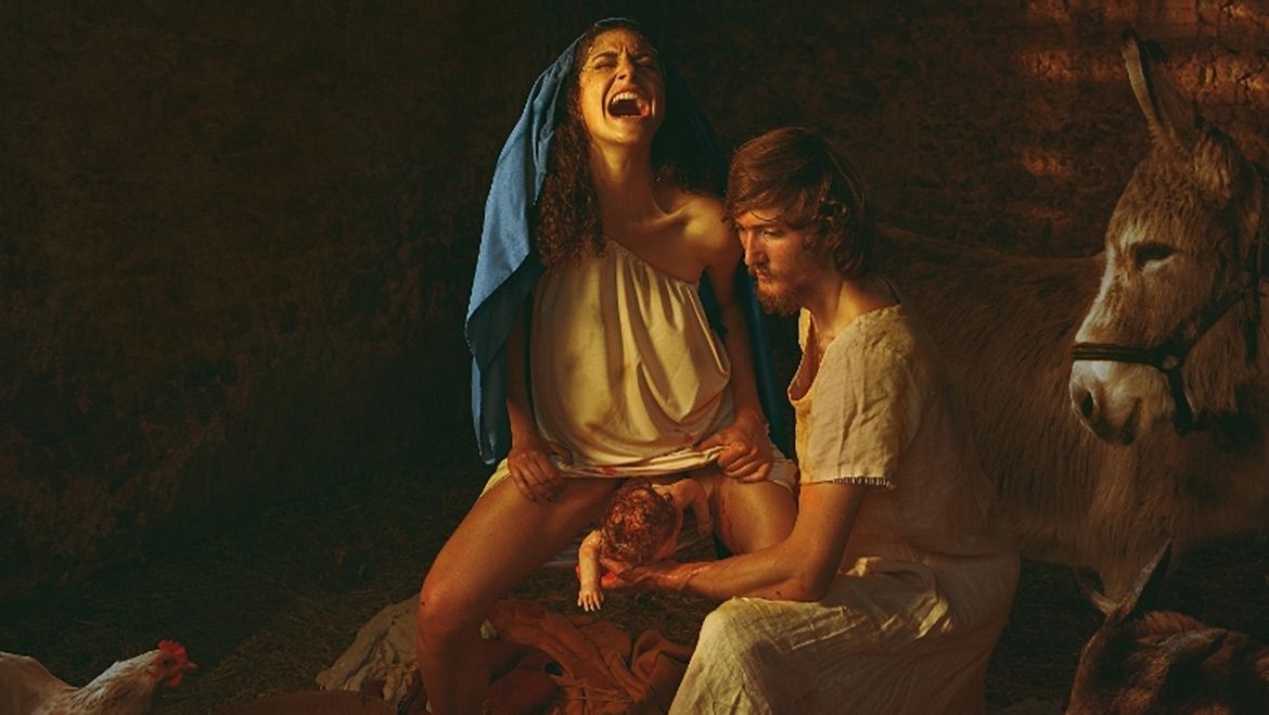 Picture of woman giving birth apparently in a stable with a person helping her and a donkey looking on