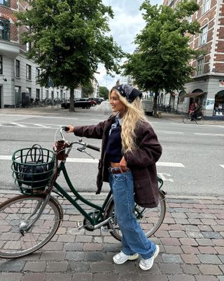 Emili wears blue wide-leg jeans, chunky New Balance sneakers, an oversize coat, bandana, belt, and navy collared shirt.