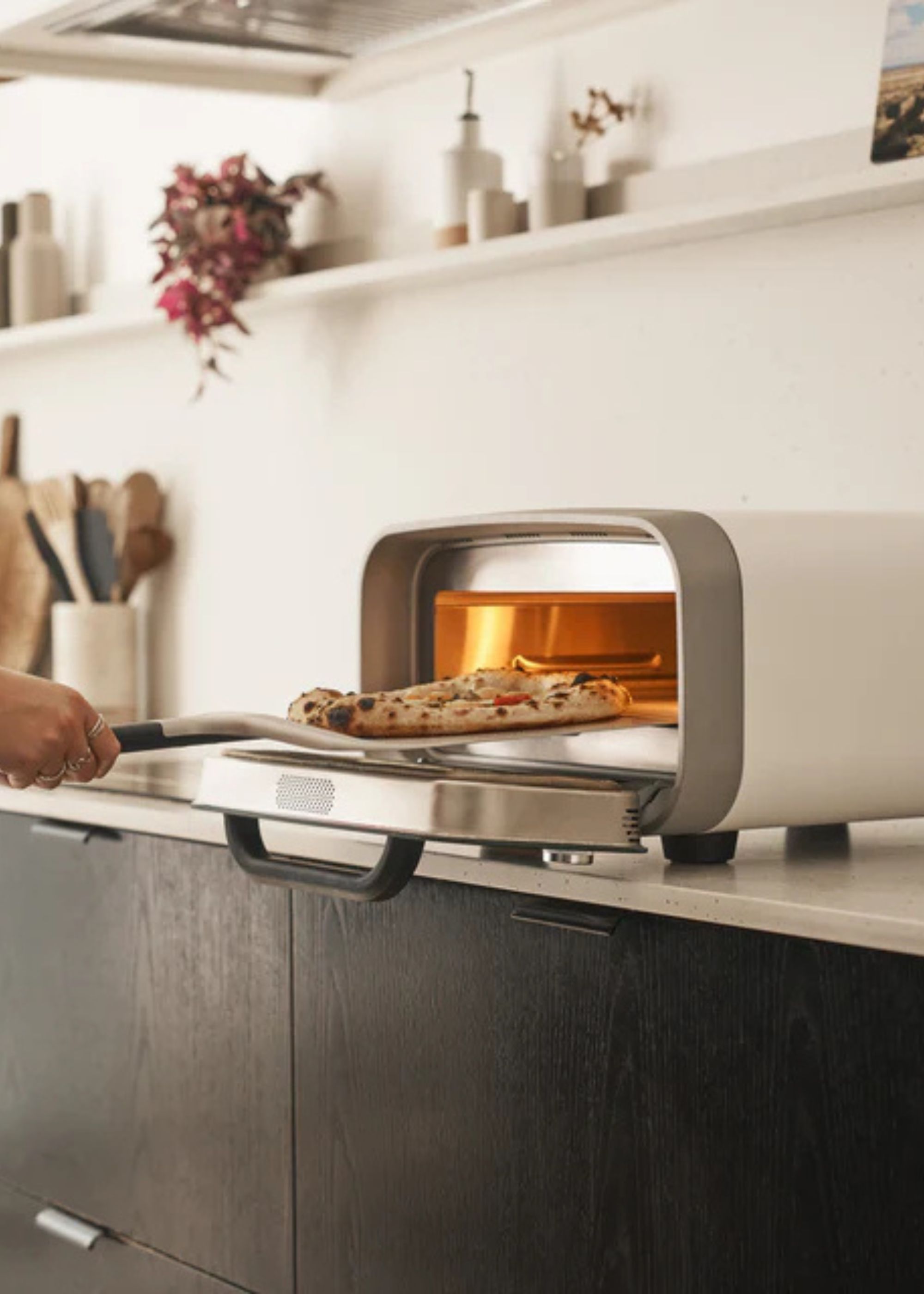 Ooni's Volt 2 indoor pizza oven on a kitchen countertop with someone taking a pizza out of the oven with a peel