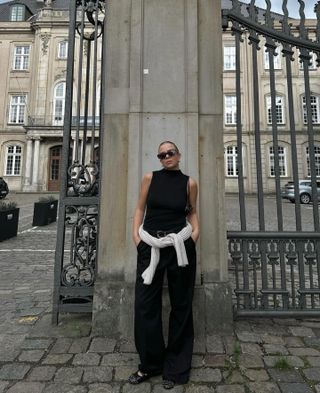 Dina Hansen wears a black turtleneck tank top, black trousers, black studded shoes, black sunglasses and a beige sweater.