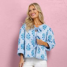 Blonde woman wearing blue Mud Pie jacket and white jeans with pink background