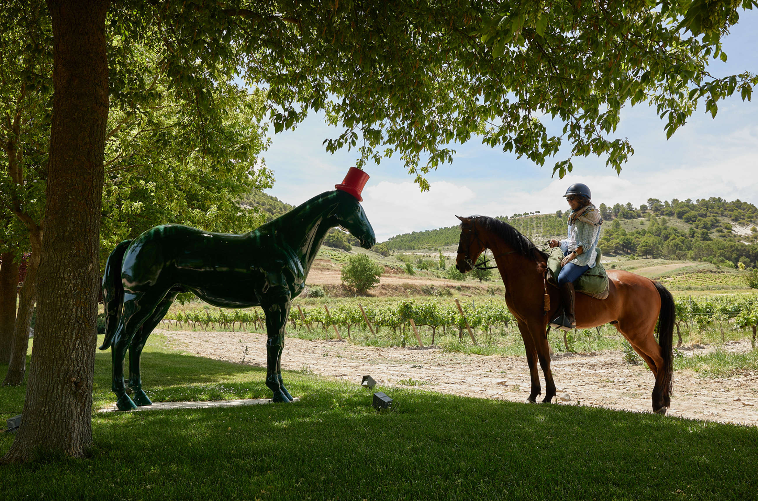 A woman on a horse, facing a large dark green statue of a horse with a red top hat on, with vines in the background