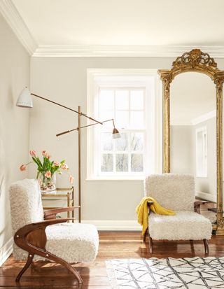 Corner of a gray-neutral living room with two white, fuzzy, and wooden accent chairs, a floor-to-ceiling ornate gold mirror and a gold and glass side table with a vase of pink tulips