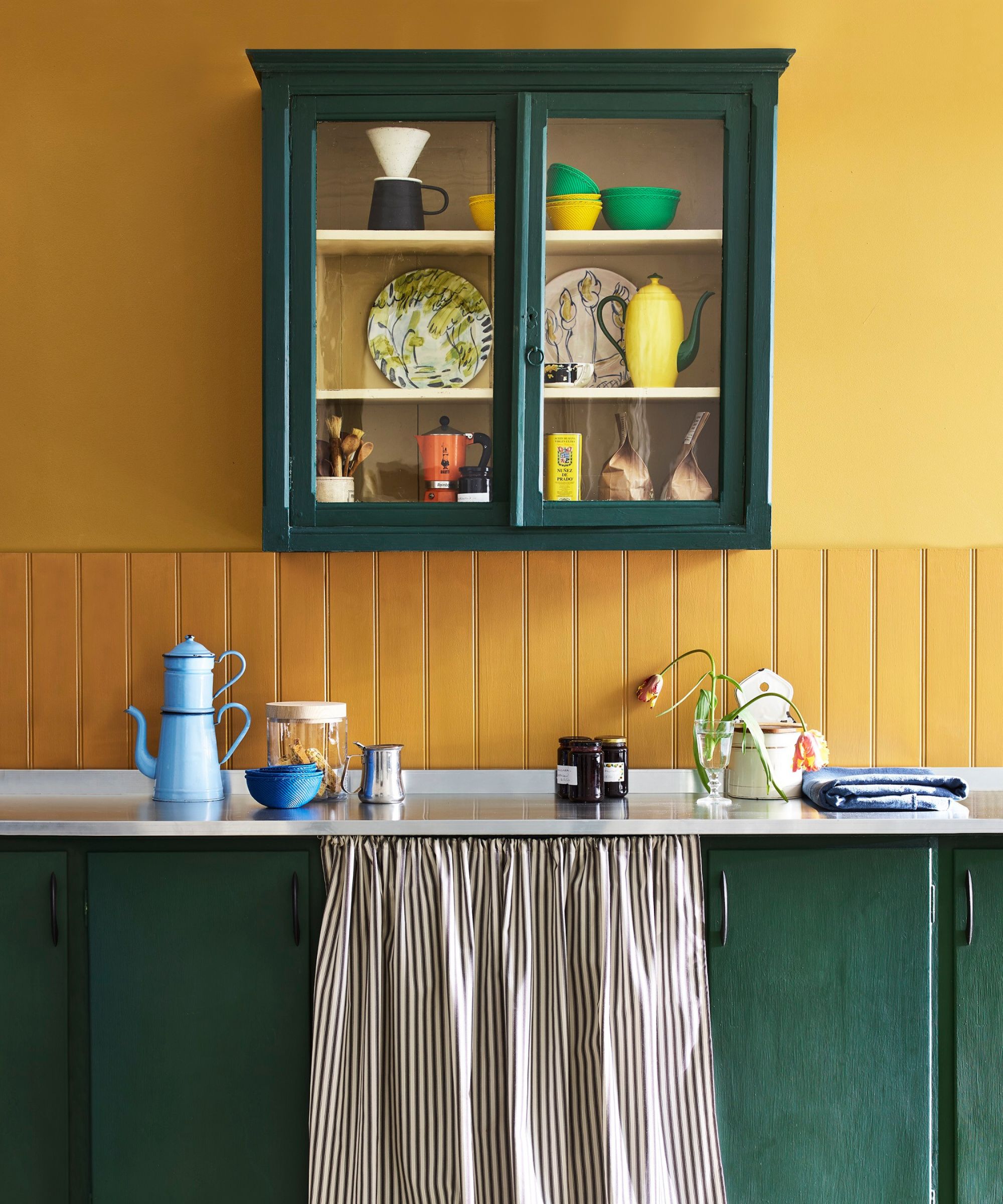 Bold kitchen with mustard yellow walls and deep green cabinetry. A glass-fronted wall cupboard displays colourful crockery and a yellow teapot. Below, a striped fabric curtain conceals storage, while a blue enamel coffee pot and jars sit neatly arranged.