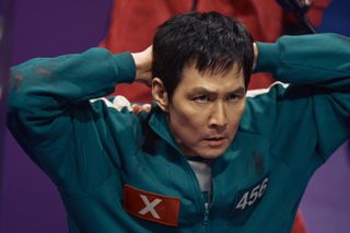 A man (Lee Jung-jae as Gi-hun) wearing a green tracksuit with a red X on one side of the chest and the number 456 on the other, crouching with his hands behind his head, in 'Squid Game' season 2.