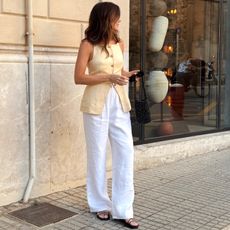 British influencer Kim Turkington poses on a sidewalk in Palma De Mallorca, Spain wearing a butter-yellow button-front vest, a black beaded mini bag, white linen pants, and flat strappy black sandals