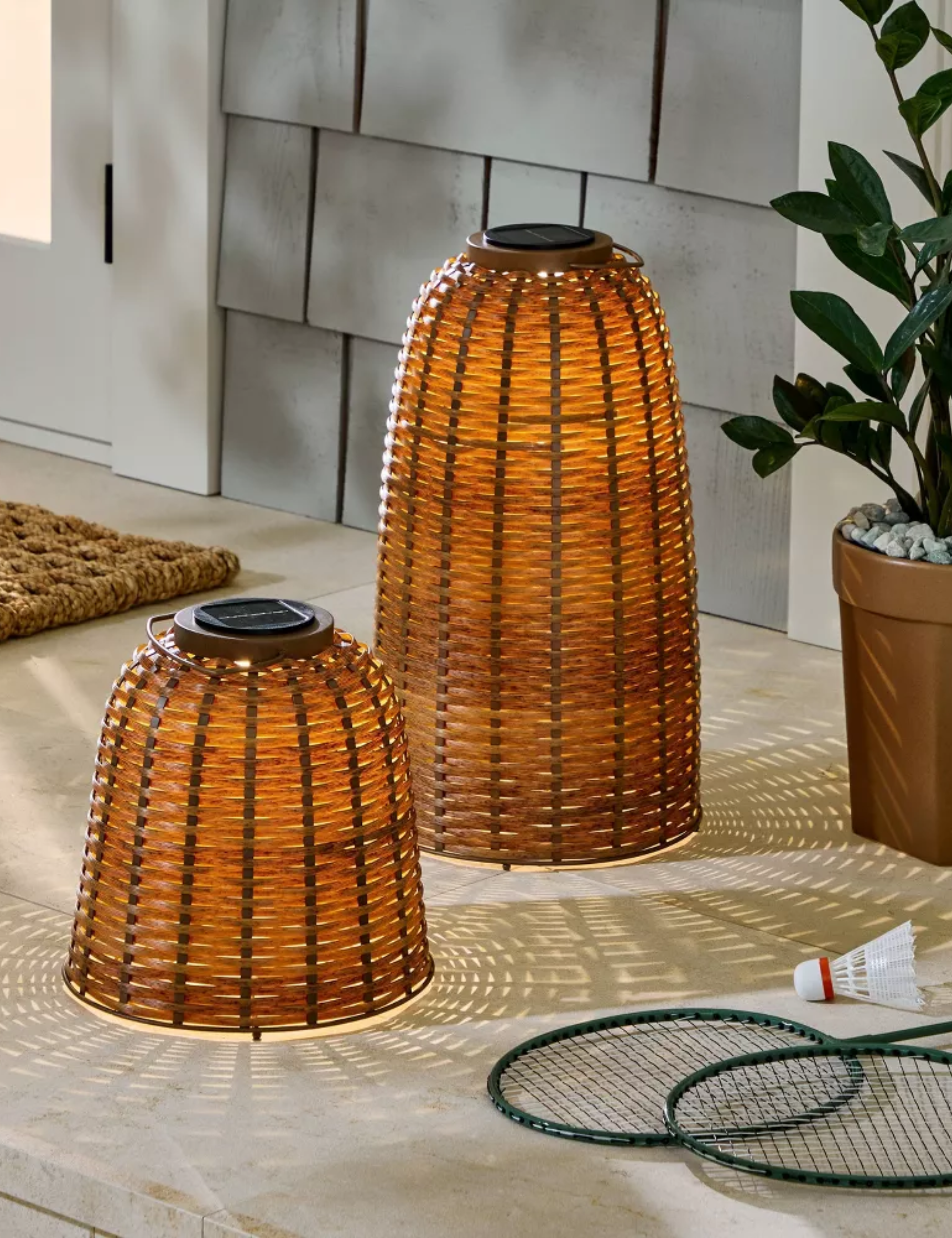 Pair of woven outdoor lanterns styled on a back porch alongside a potted plant and badminton accessories.