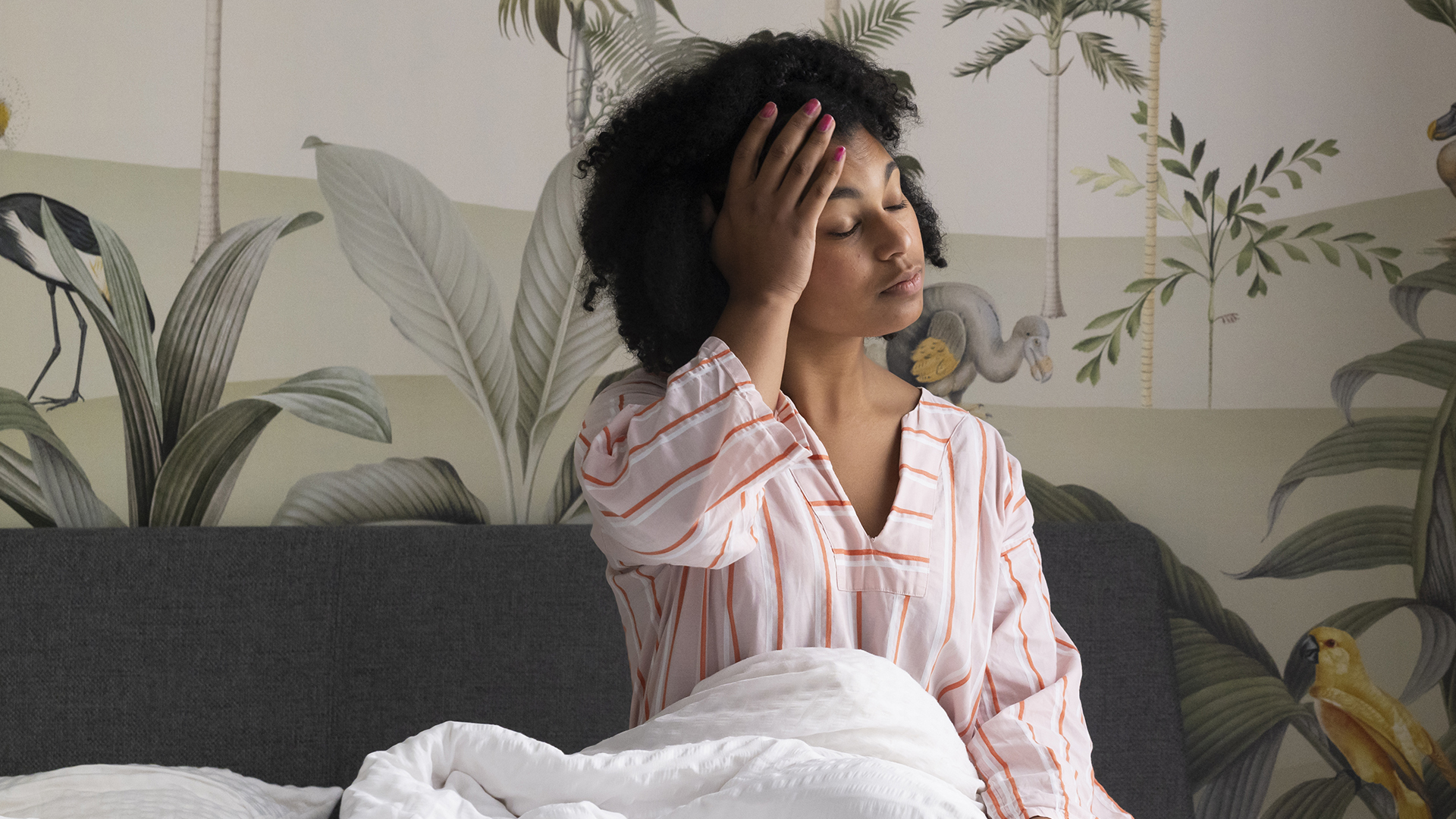 A woman sits up in her bed with her hand on her head looking exhausted and like she hasn't had much sleep
