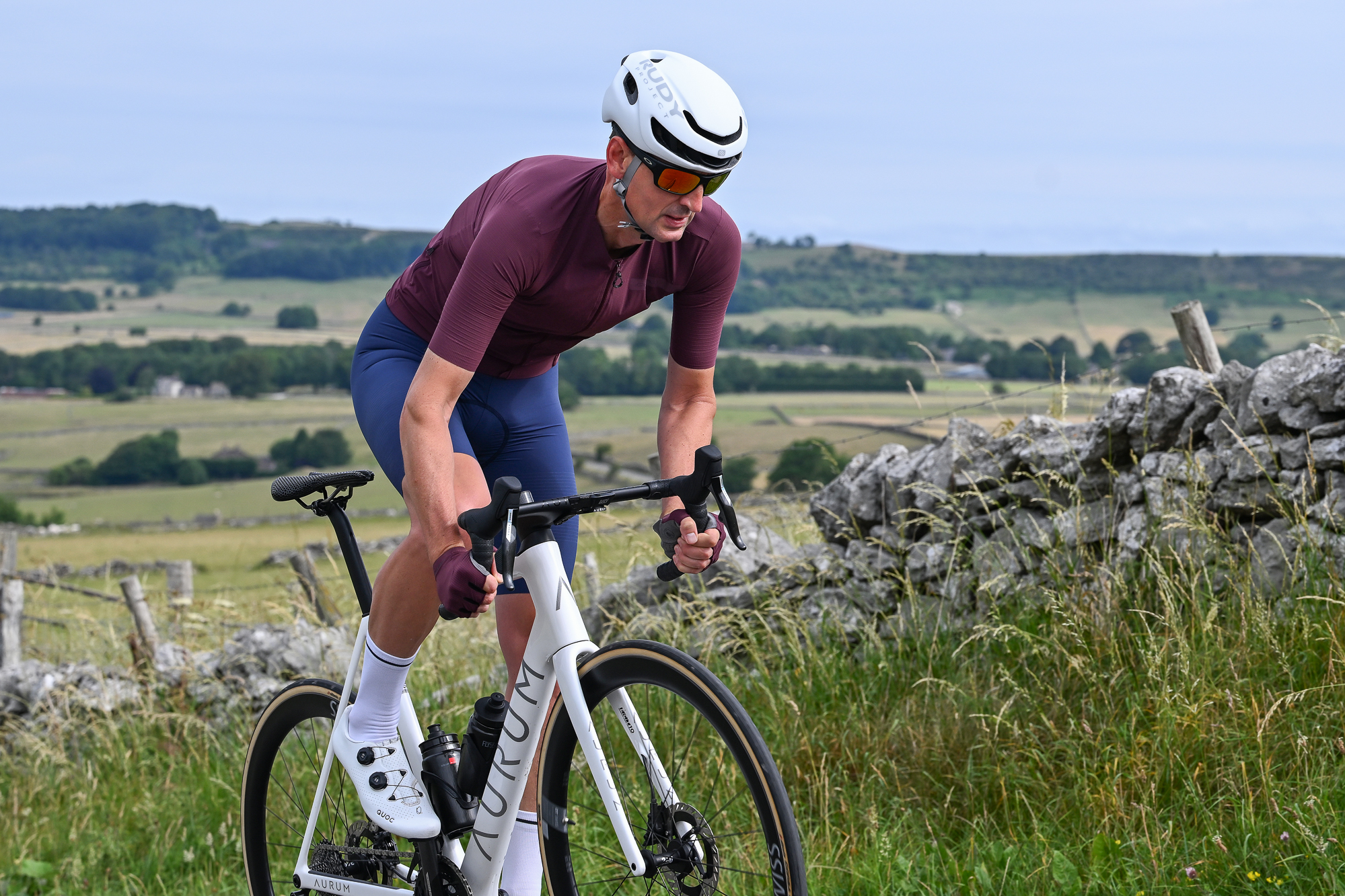 Man wearing a purple jersey, blue cycling shorts and a white helmet riding a white bike uphill