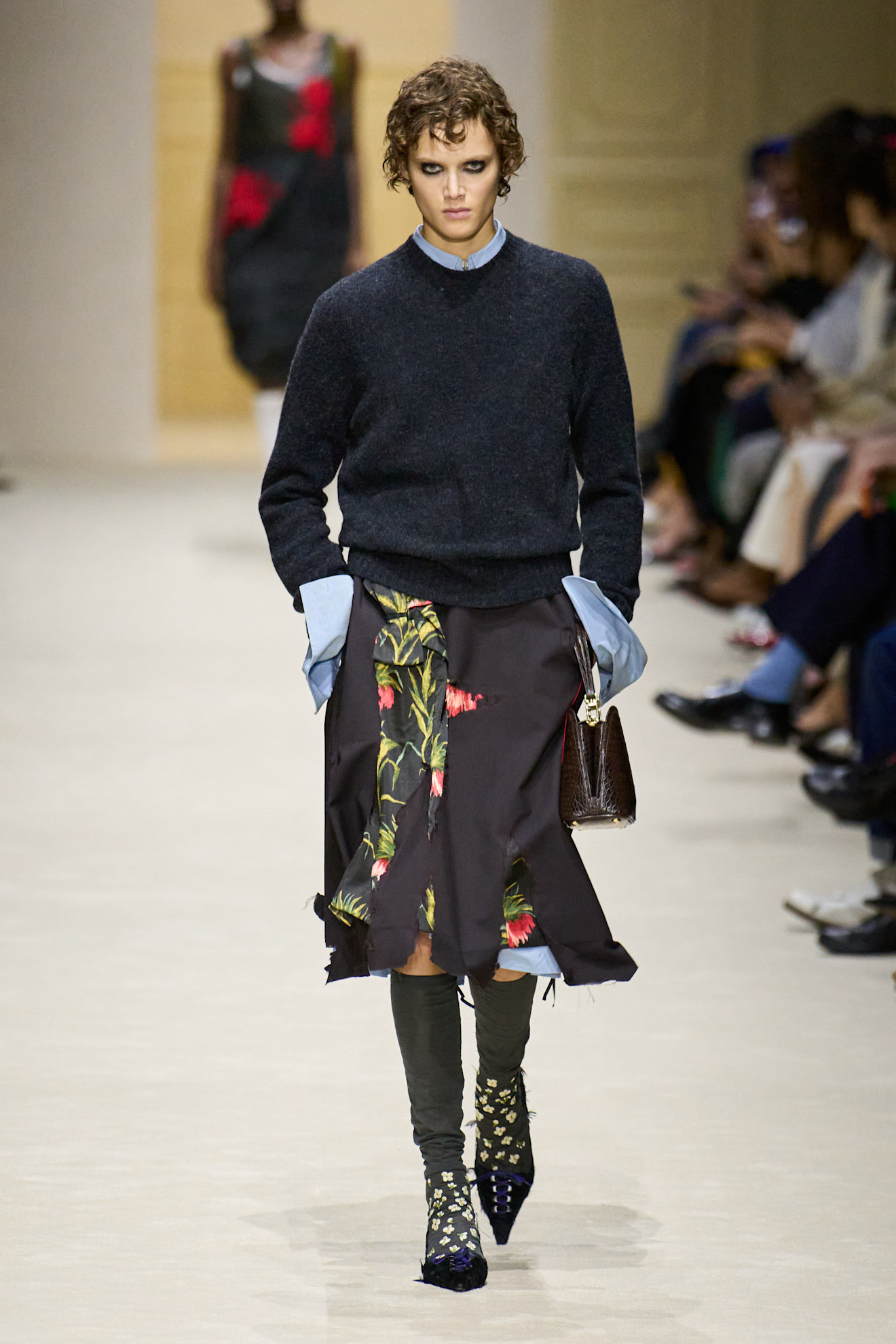 A model at the F/W 26 Prada show wearing a slasher skirt with knee-high socks, pumps, and a sweater.