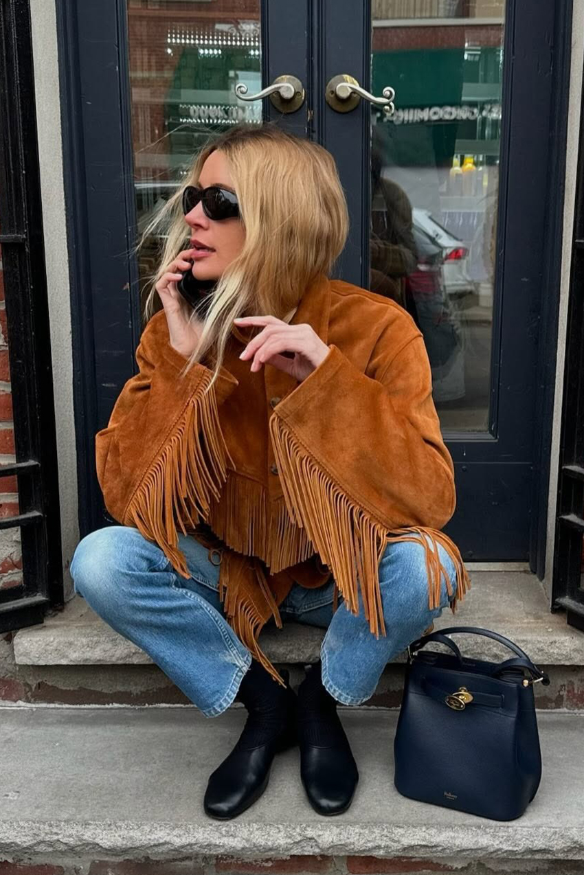 a woman wearing a fringe jacket, blue jeans, black boots, and a black bag